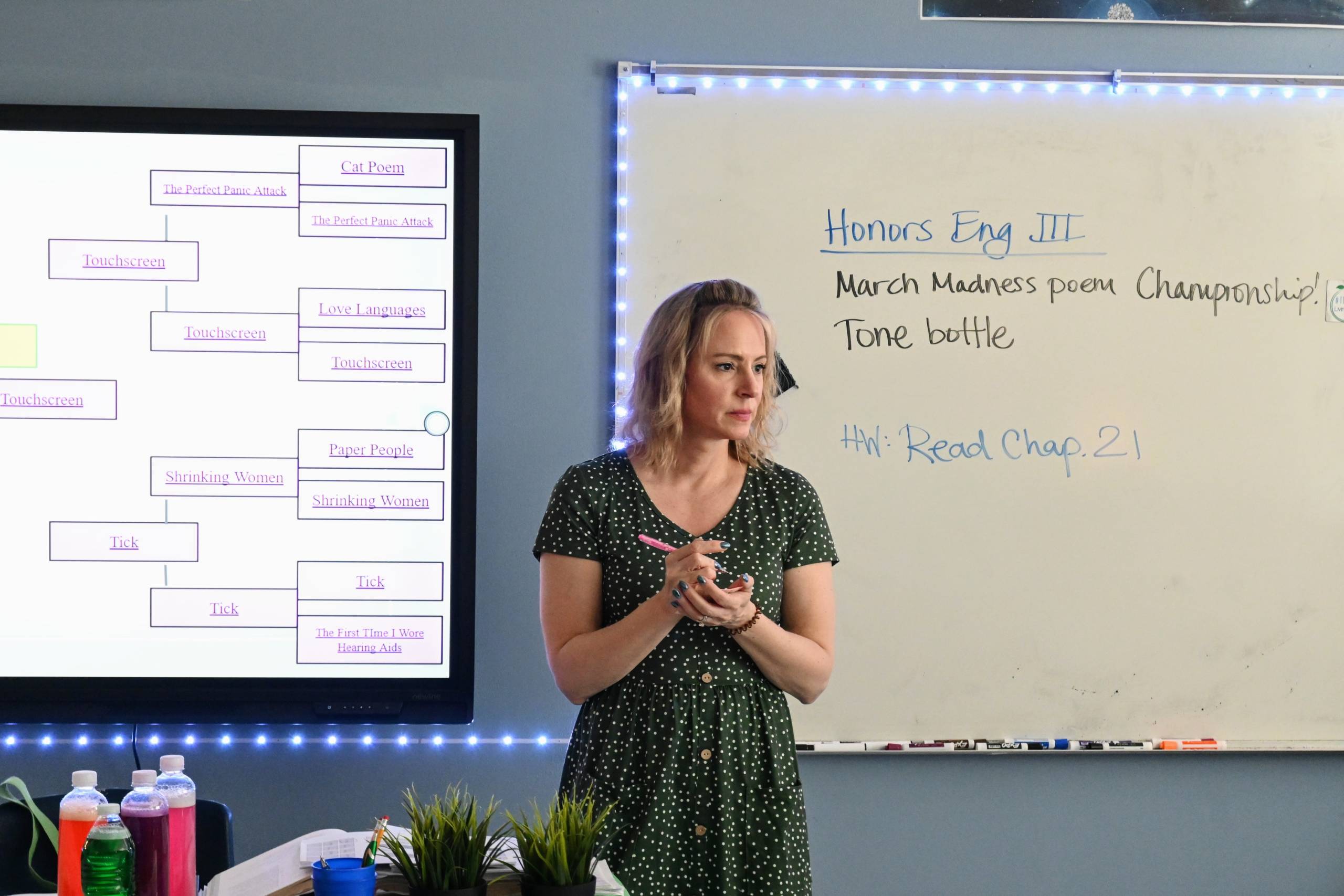 A white woman in a polka dot dress holds a pen and pad of sticky notes. She stands in front of a white board.