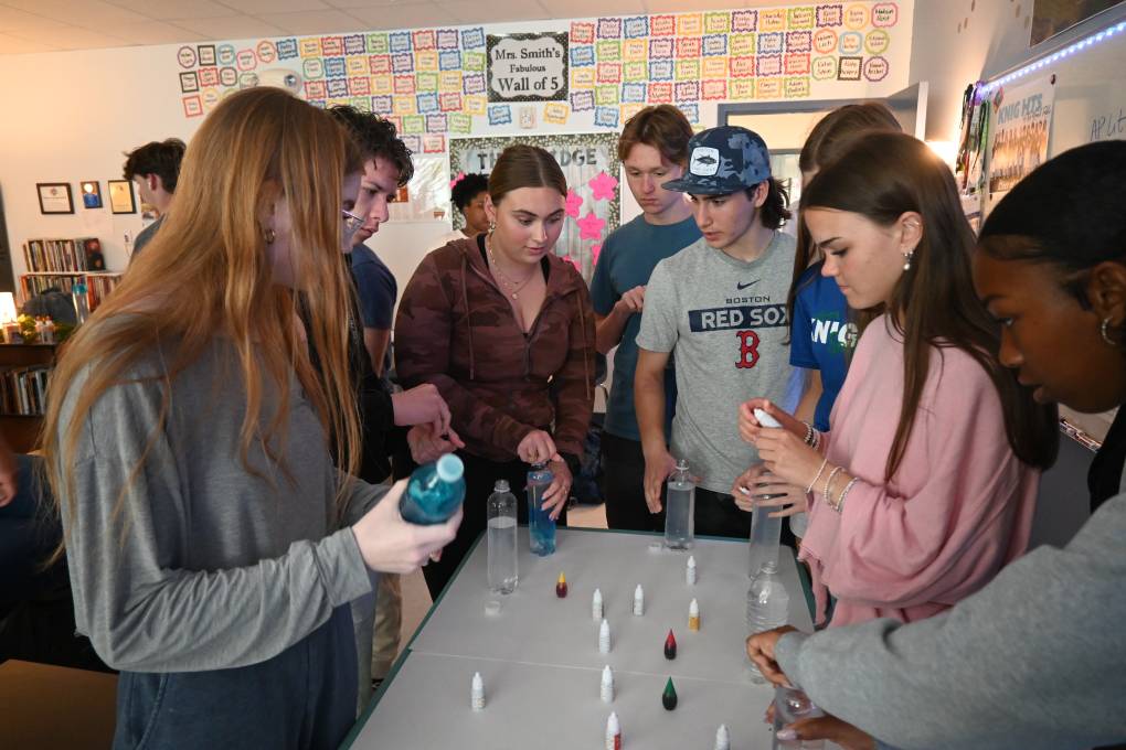 A group of high school students gathered around a classroom table holding plastic bottles. Small containers of food color are arranged on the table.