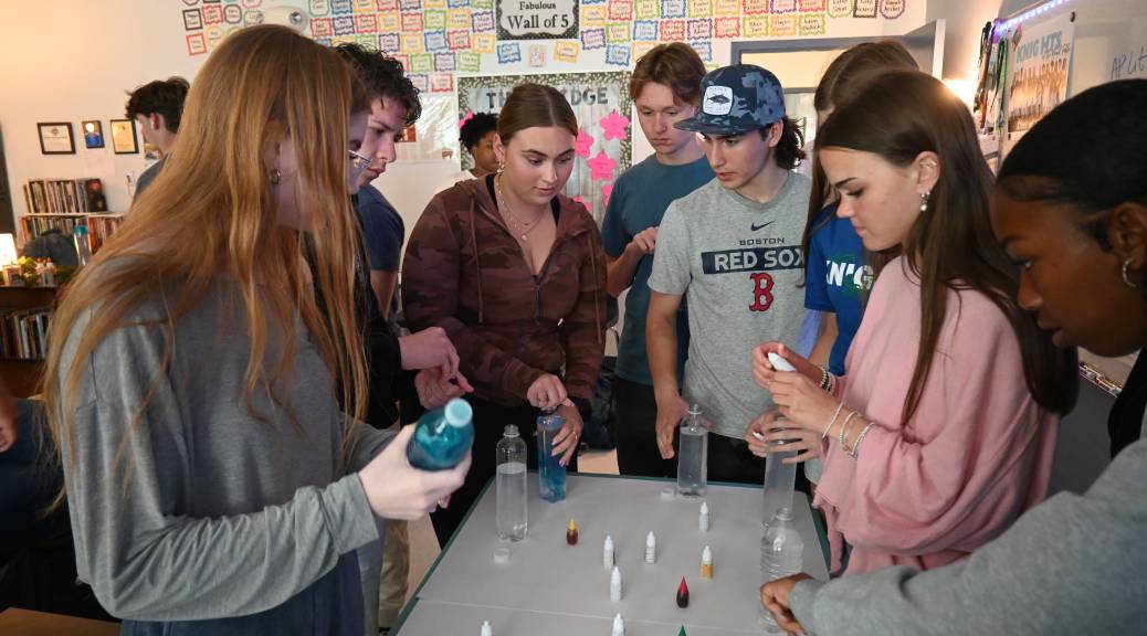 A group of high school students gathered around a classroom table holding plastic bottles. Small containers of food color are arranged on the table.