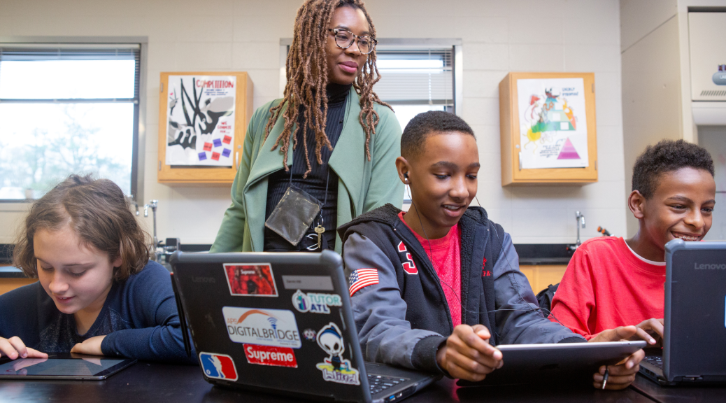 An adult overlooks three students shoulders as they work on computers.