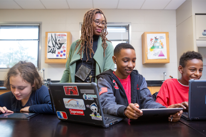 An adult overlooks three students shoulders as they work on computers.