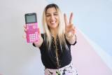 A smiling woman with blonde hair and dark roots wearing a black shirt and patterned pants holds out a calculator and peace fingers.