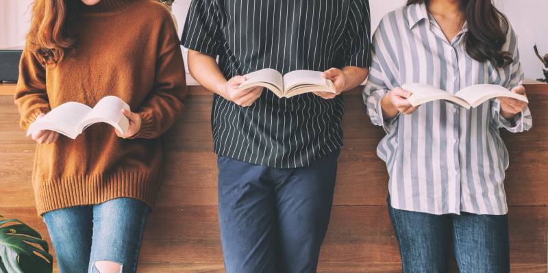 Three people lean against a wall, each holding an open book. The people ar only shown from shoulders down to knees.