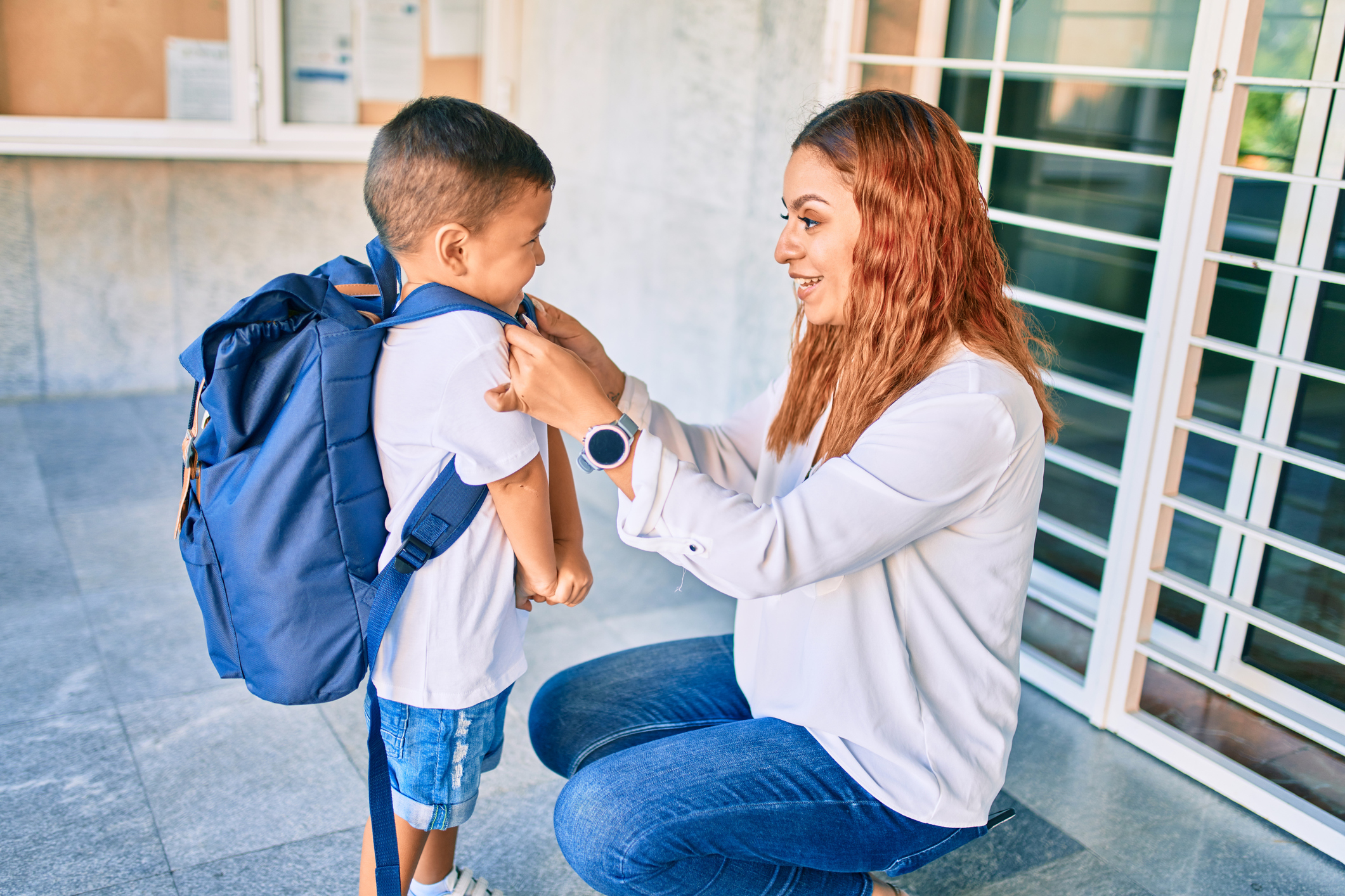 Adult woman kneels in front of a little boy wearing a backpack at a school entrance