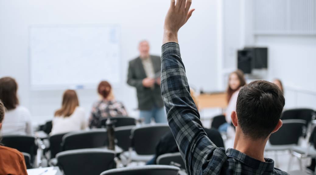 male student raises his hand at the back of a college classroom