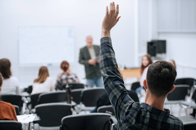 male student raises his hand at the back of a college classroom