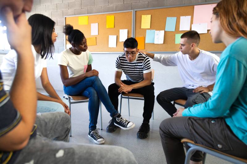 Group of teenagers in sit in a circle on chairs looking toward one young man whose eyes look to the floor. The young man next him rest his hand on the other's shoulder.