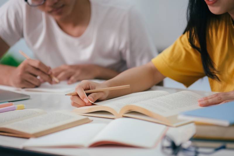 One student in a white tshirt and another student in a yellow tshirt sit at.a desk with pencils in hand and several books laying open on the desk.