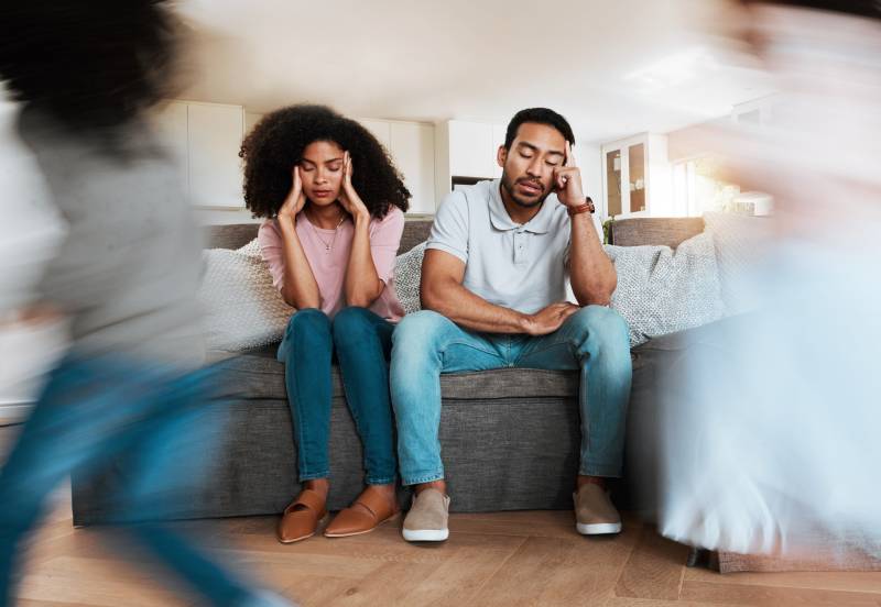 Two parents sit on a couch with heads on their head in stressed expression. Two children run in front of them with blurred focus