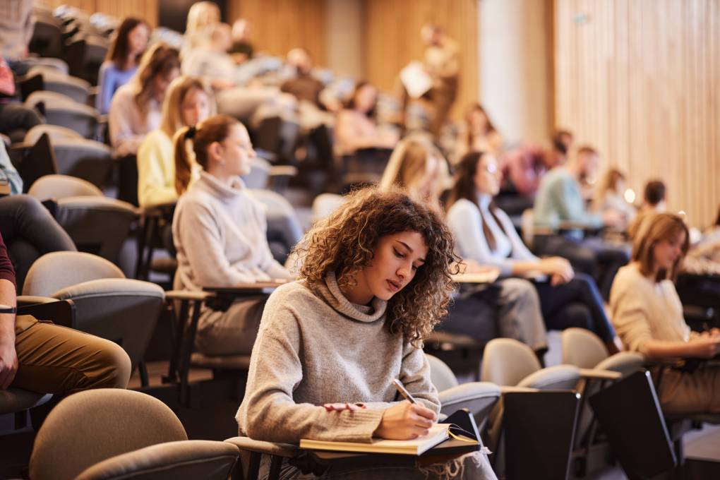 Female college student writing an exam during a class at lecture hall. Her classmate are in the background.