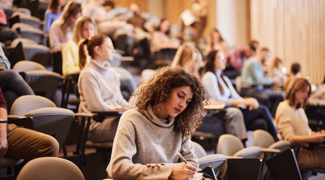 Female college student writing an exam during a class at lecture hall. Her classmate are in the background.