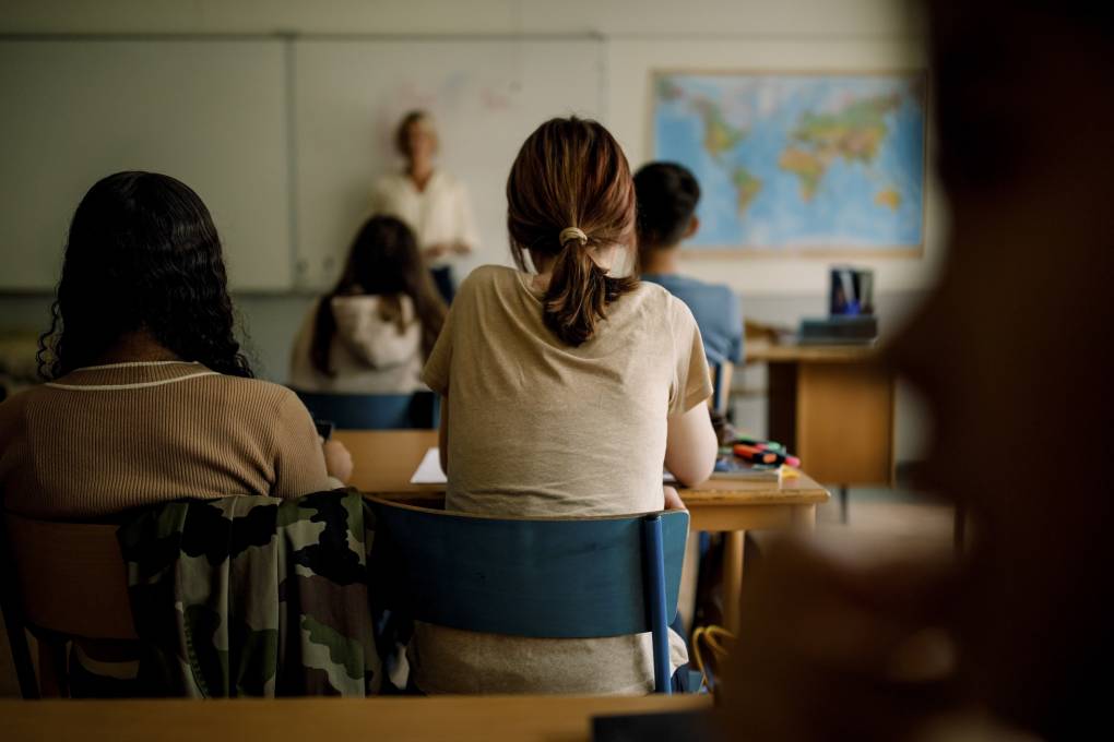 View of the backs of students who are listening to their teacher at the front of the classroom