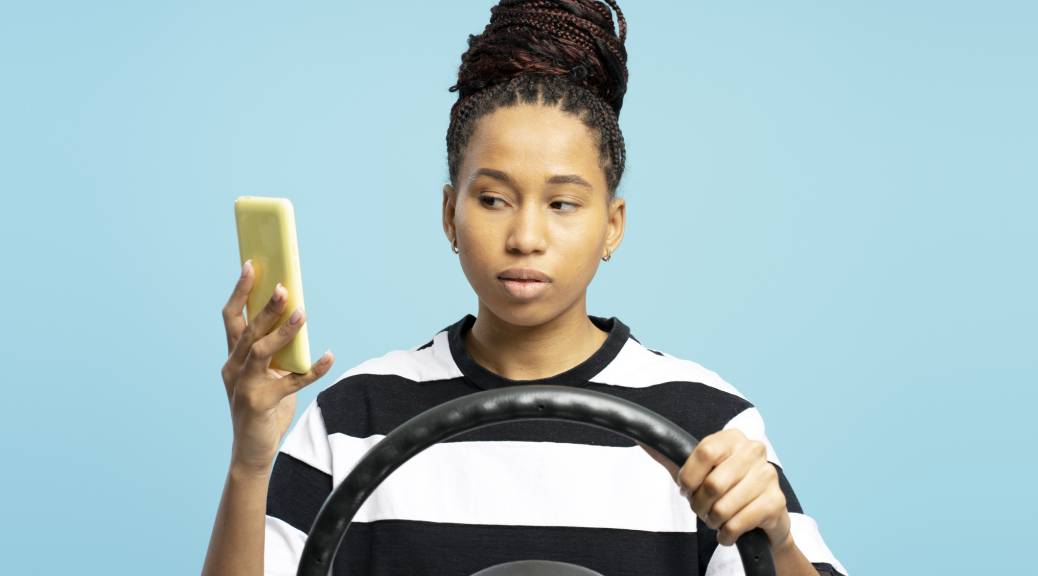Studio portrait of a young woman gripping a steering wheel while engaging with smartphone