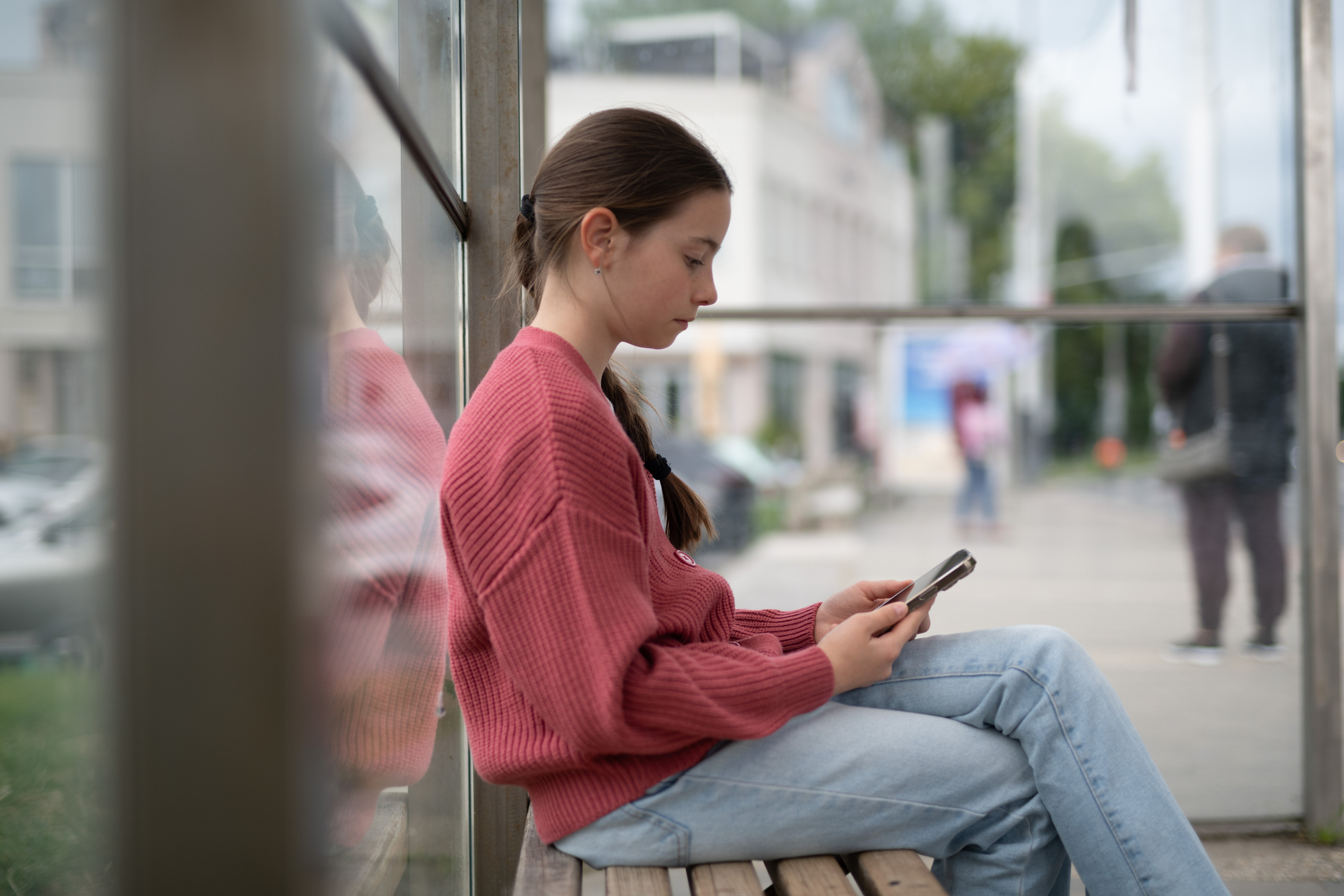 Young girl sitting at bus stop, scrolling through phone.