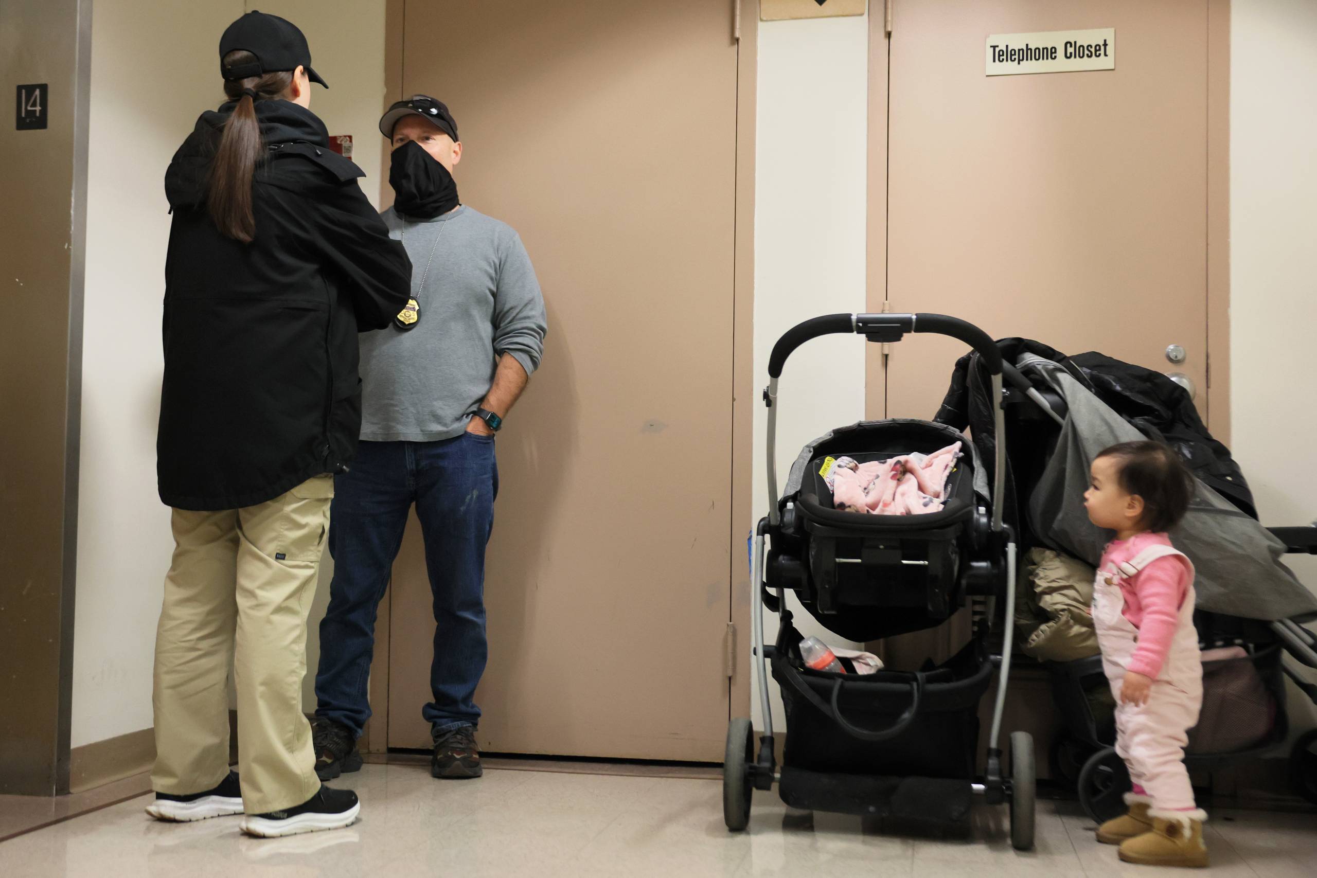 A masked adult talks to another adult as a toddler stands next to a stroller.