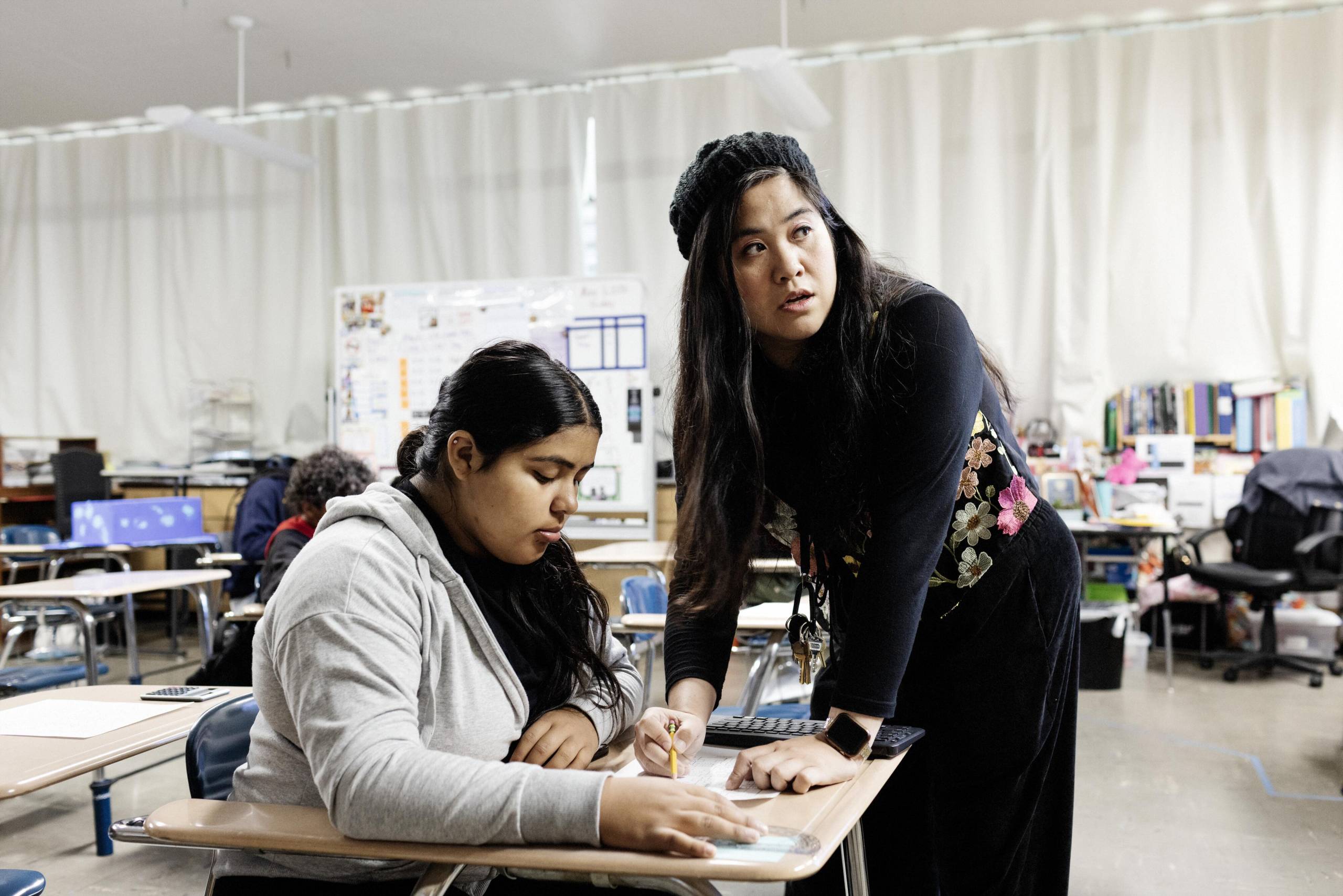 A teacher helps a student who is seated at her desk.