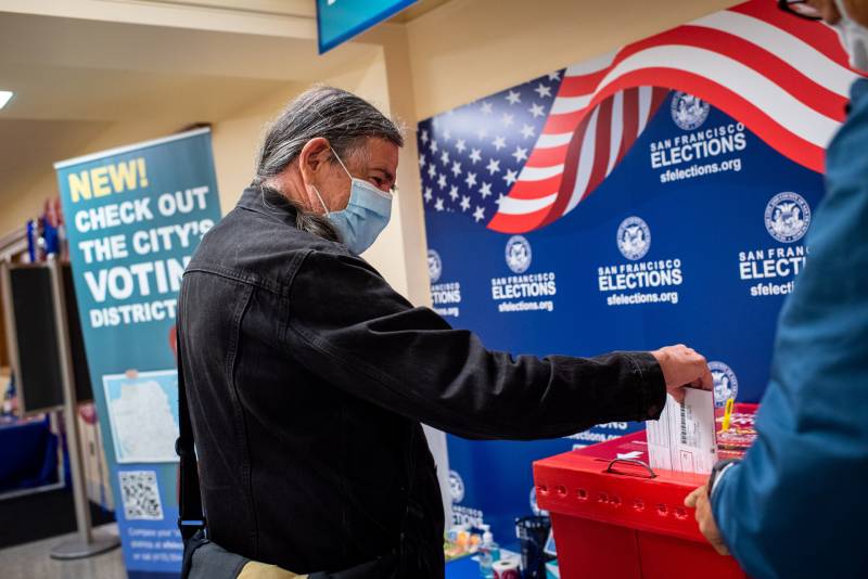 A man with a mask drops his ballot into a ballot box.