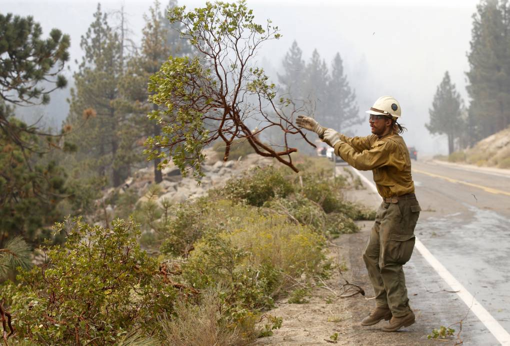 A man in a hard hat, wearing heavy brown pants, a gold shirt and work gloves, is on the side of a two-lane highway with smoky air obscuring trees in the distance. He has just tossed a large branch toward a pile of branches as part of clearing vegetation during a controlled burn.