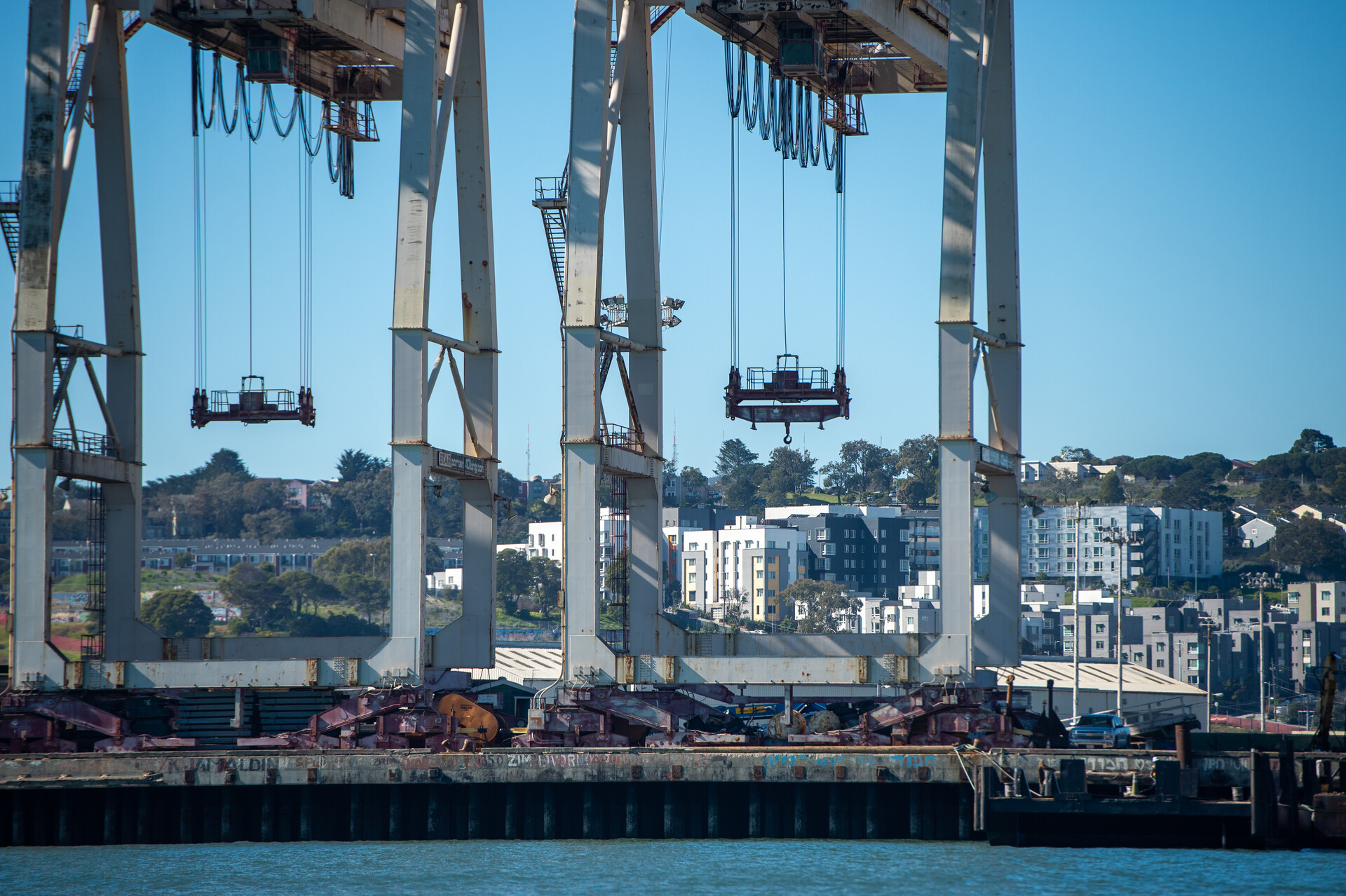 A sliver of greenish blue baywater in the foreground. Giant silver metal cranes sit upon a rock levee. Behind the cranes is an apartment laden hill. The mostly white apartments appear to be multiple stories.