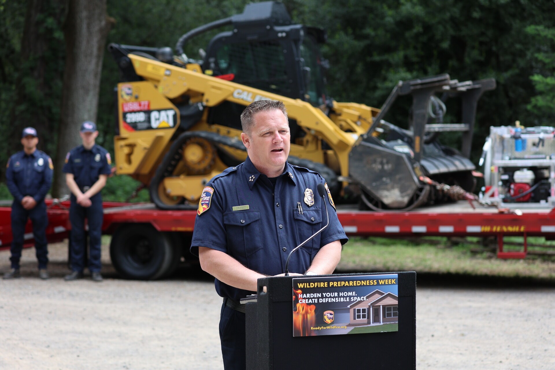 A man in a blue uniform stands at a podium in front of a bulldozer.