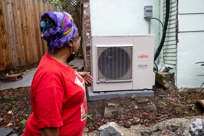 A woman in a red shirt points to the metal heat pump that sits along the outside of her home.