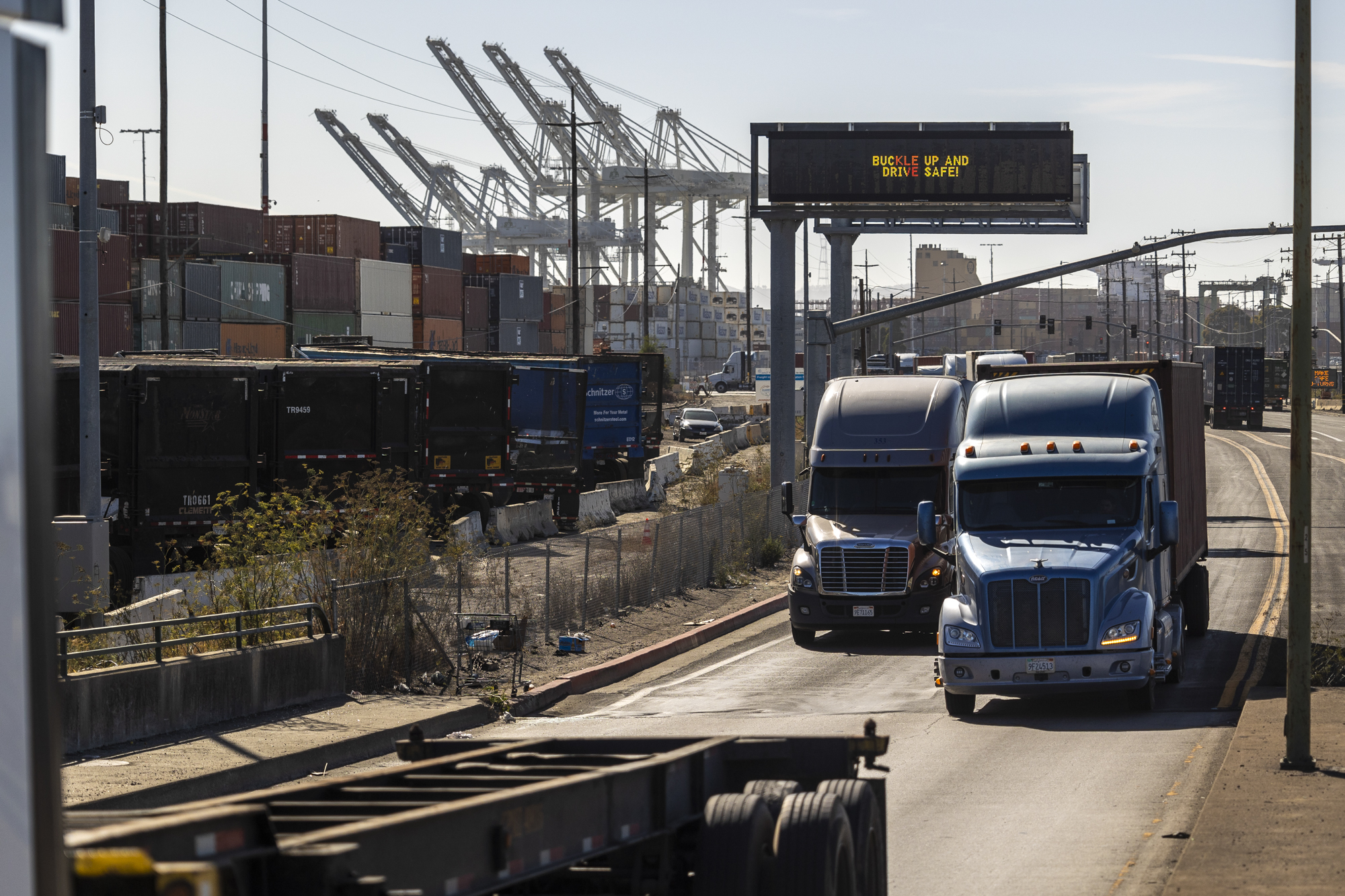 Several trucks drive through an industrial area with cranes in the background.