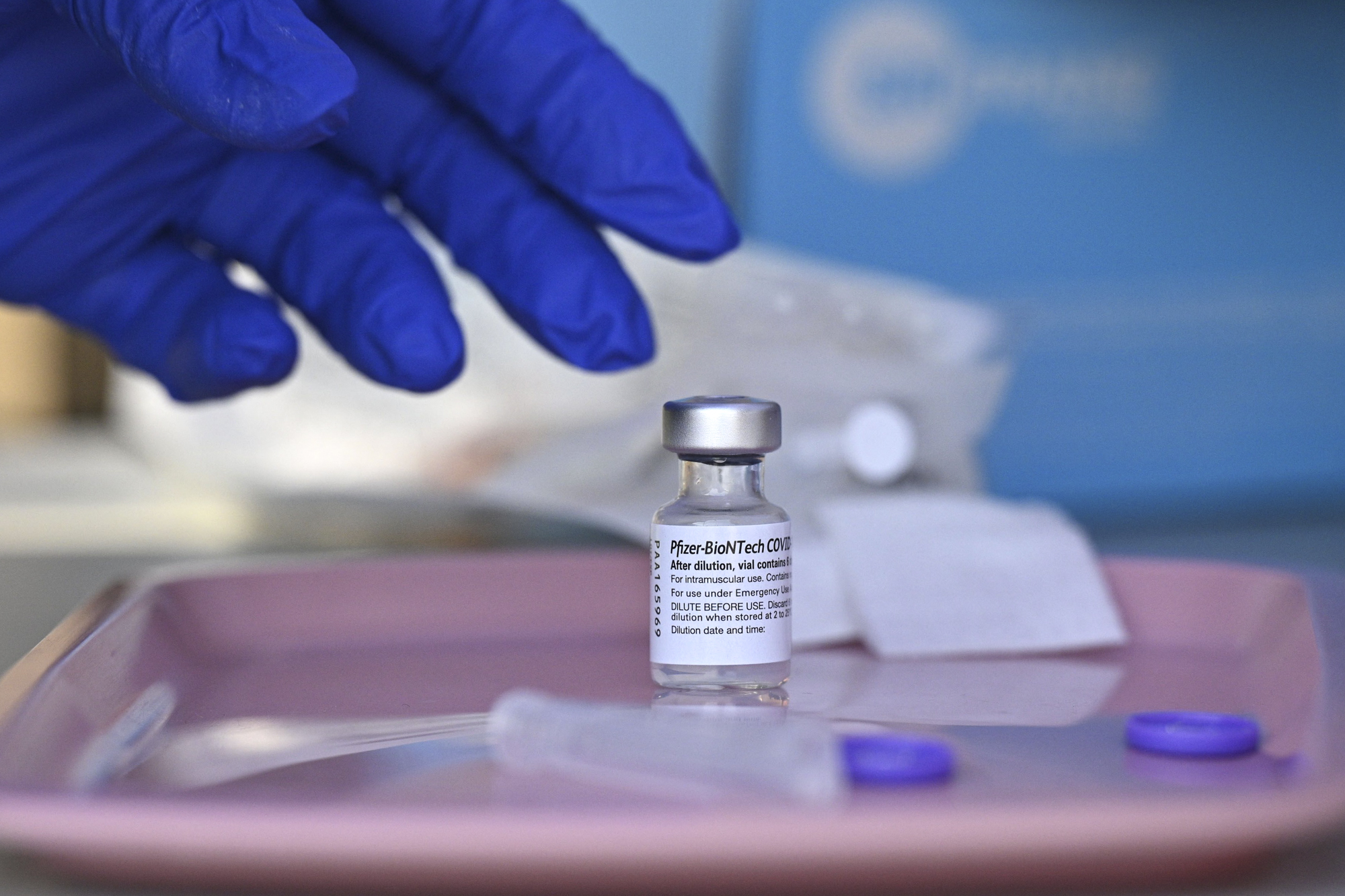 A gloved hand reaches toward a small vial on a pink tray.