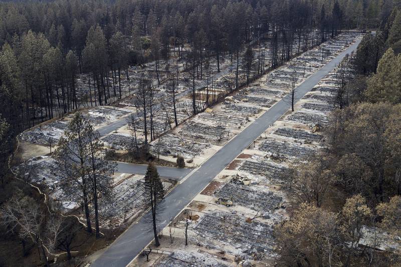 A stretch of empty land filled with the charred remains of mobile homes and surrounded by trees, some of which are visibly burnt.