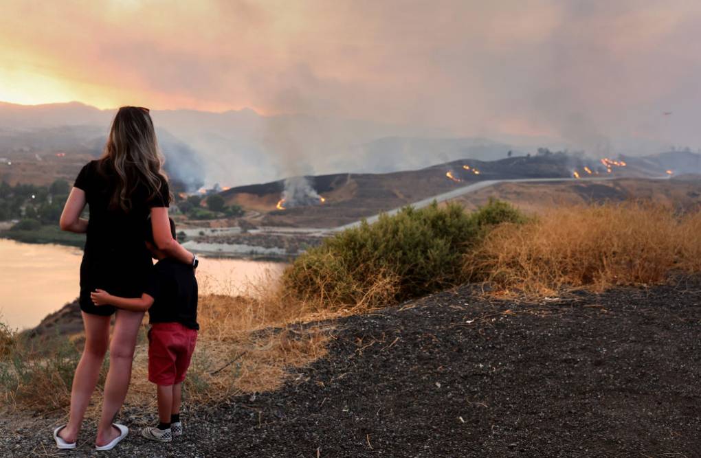 Seen from behind, a woman embraces a young child, as the two watch a fire in the distance.