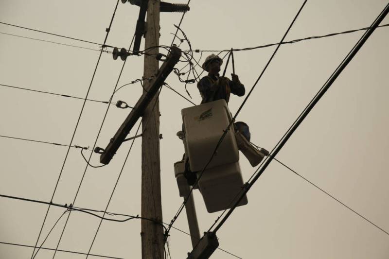 A worker in a cherry picker cuts a power line, with a gray sky in the background.