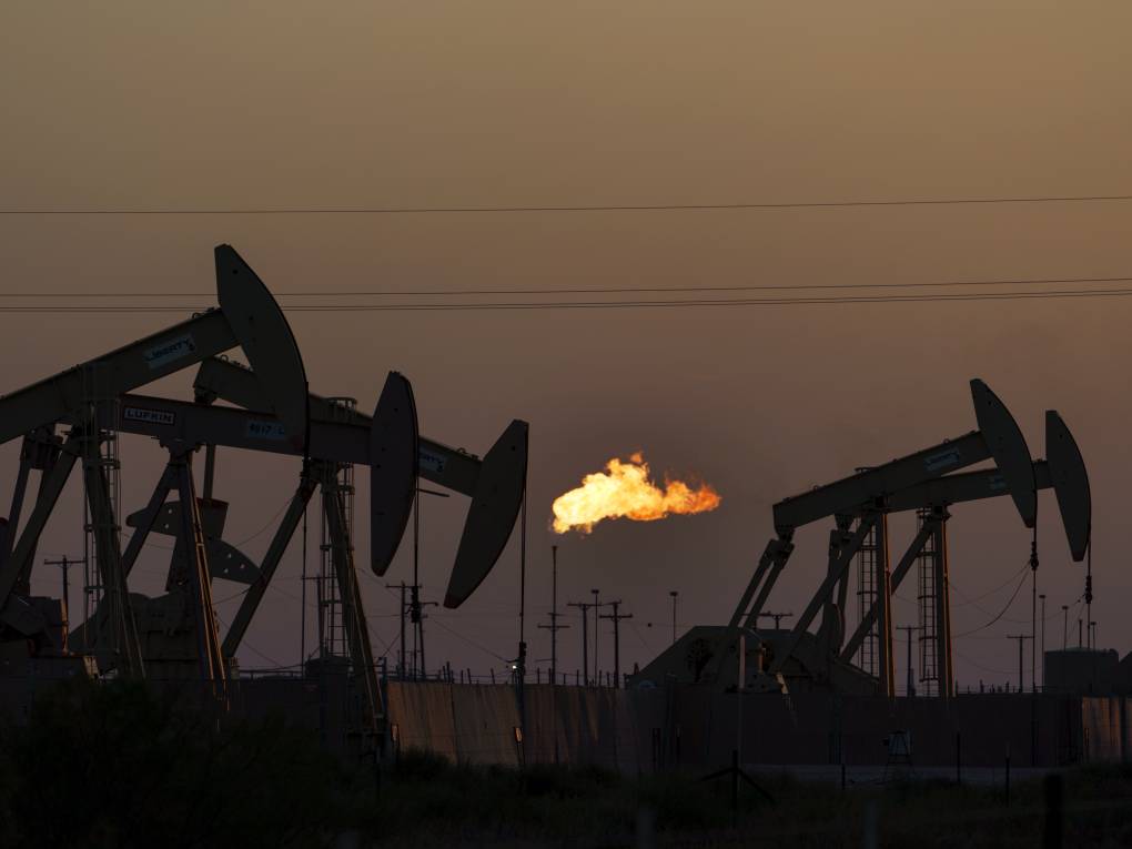 Oil pumps work in the foreground as an open flame burns in the distance beneath a smoky orange sky.
