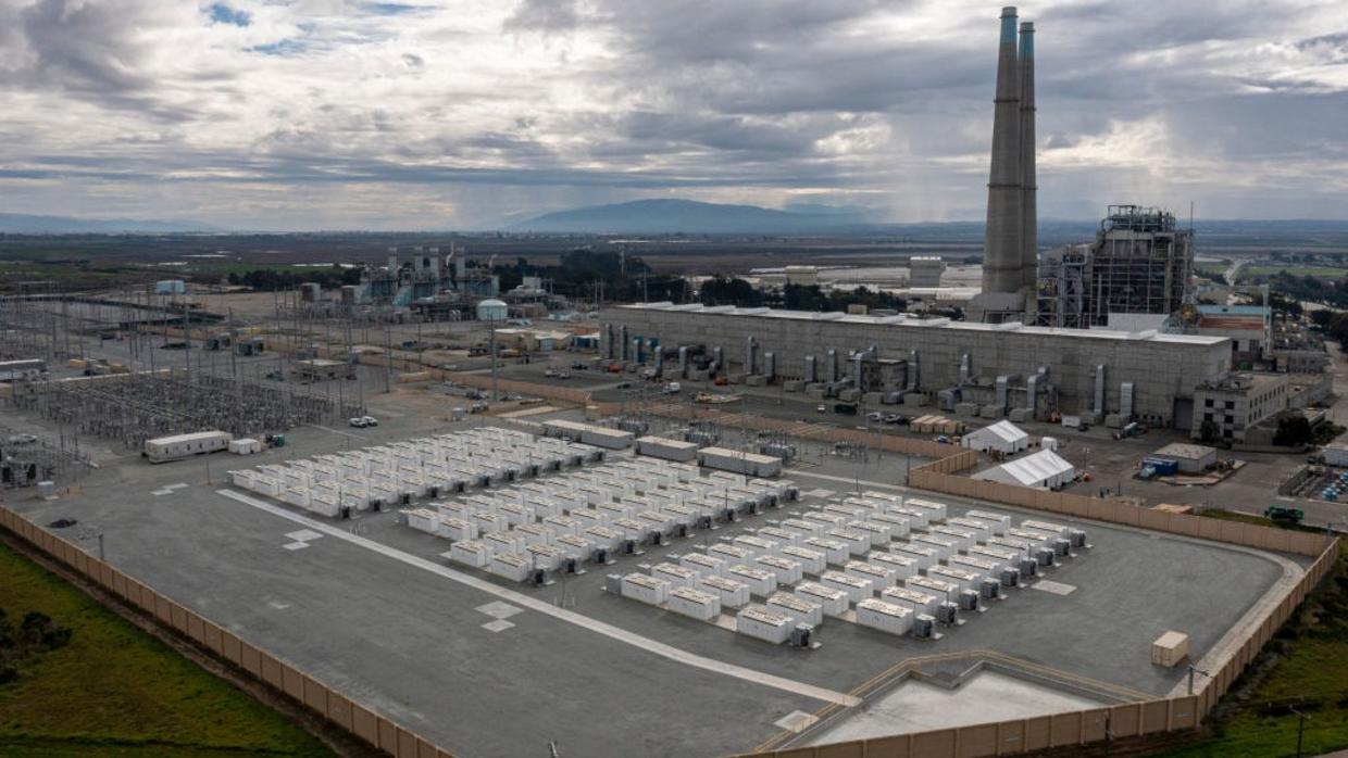 A large outdoor battery-storage facility next to a power plant with a large smokestack.