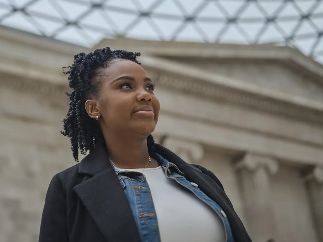 A young African American woman with white t-shirt and blazer looks away from the camera with a slight smile.