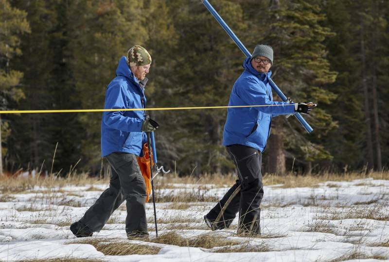 Two people in blue jackets walk through a snowy field holding equipment.