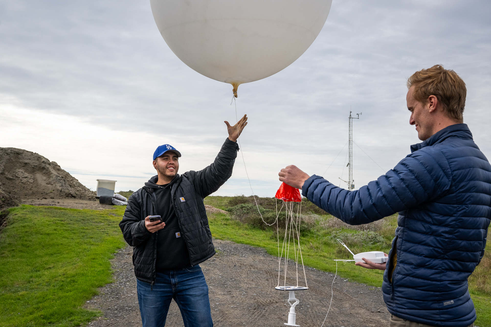 A person wearing a black jacket and blue LA Dodgers hat opens their hand to let go of a string on a large white balloon while another person in a blue jacket holds a red parachute attached to the balloon.