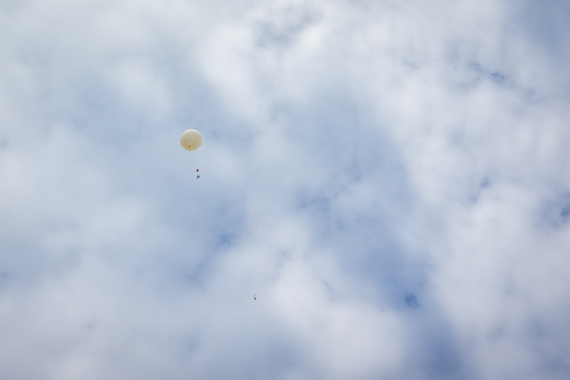 A white balloon with a string hanging from it floats in the sky with clouds behind it.