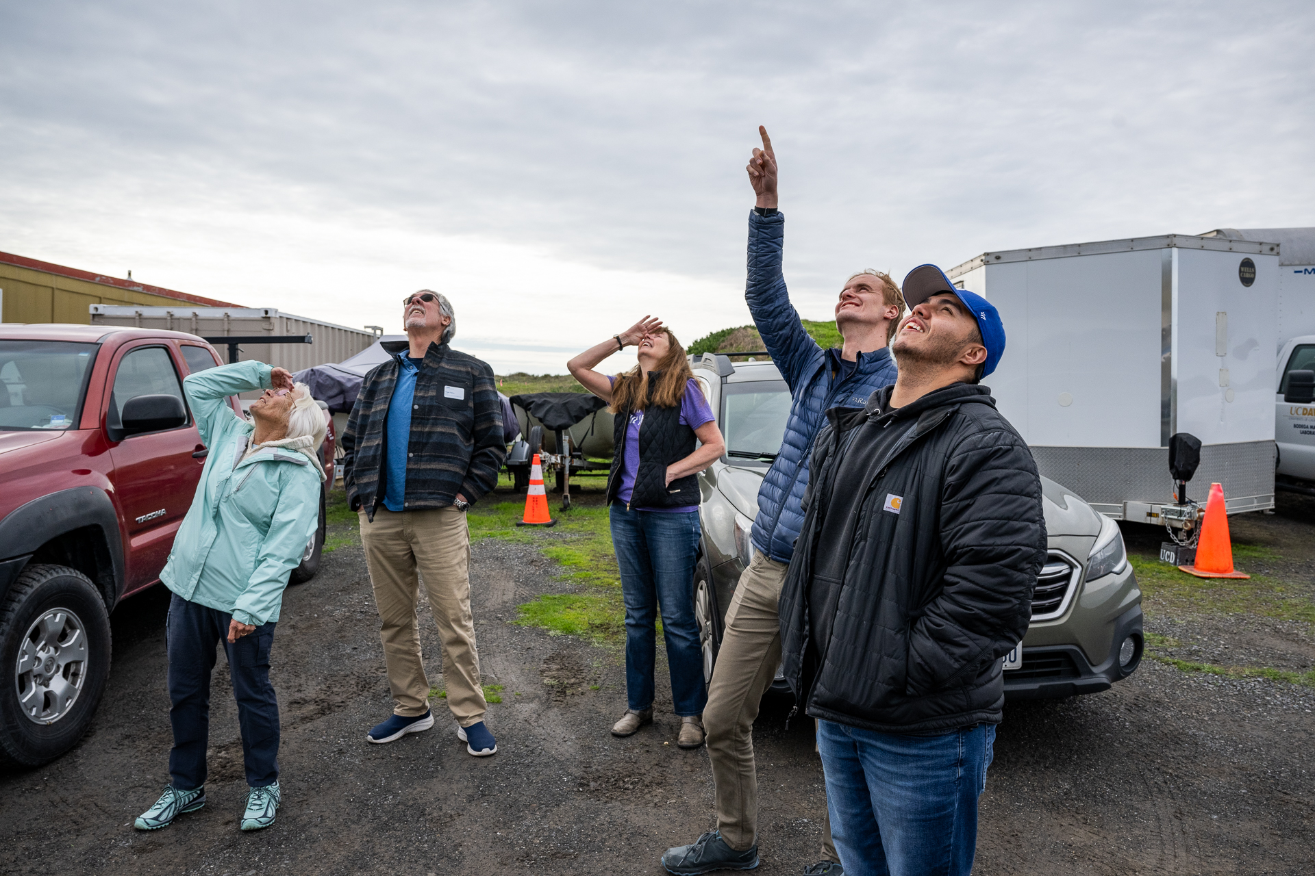 Five people look up toward an overcast sky with vehicles behind them.
