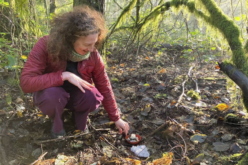 A person with long hair kneels near some small objects on the ground in a wooded area.
