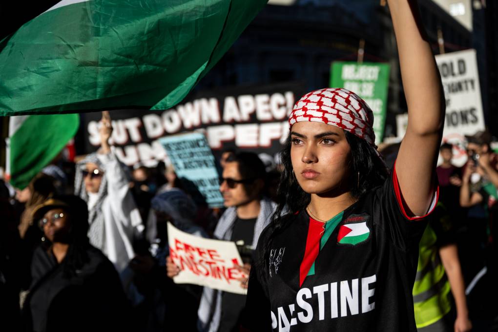 A woman with a black shirt that says "Free Palestine" holds her left hand up in the air in a fist.