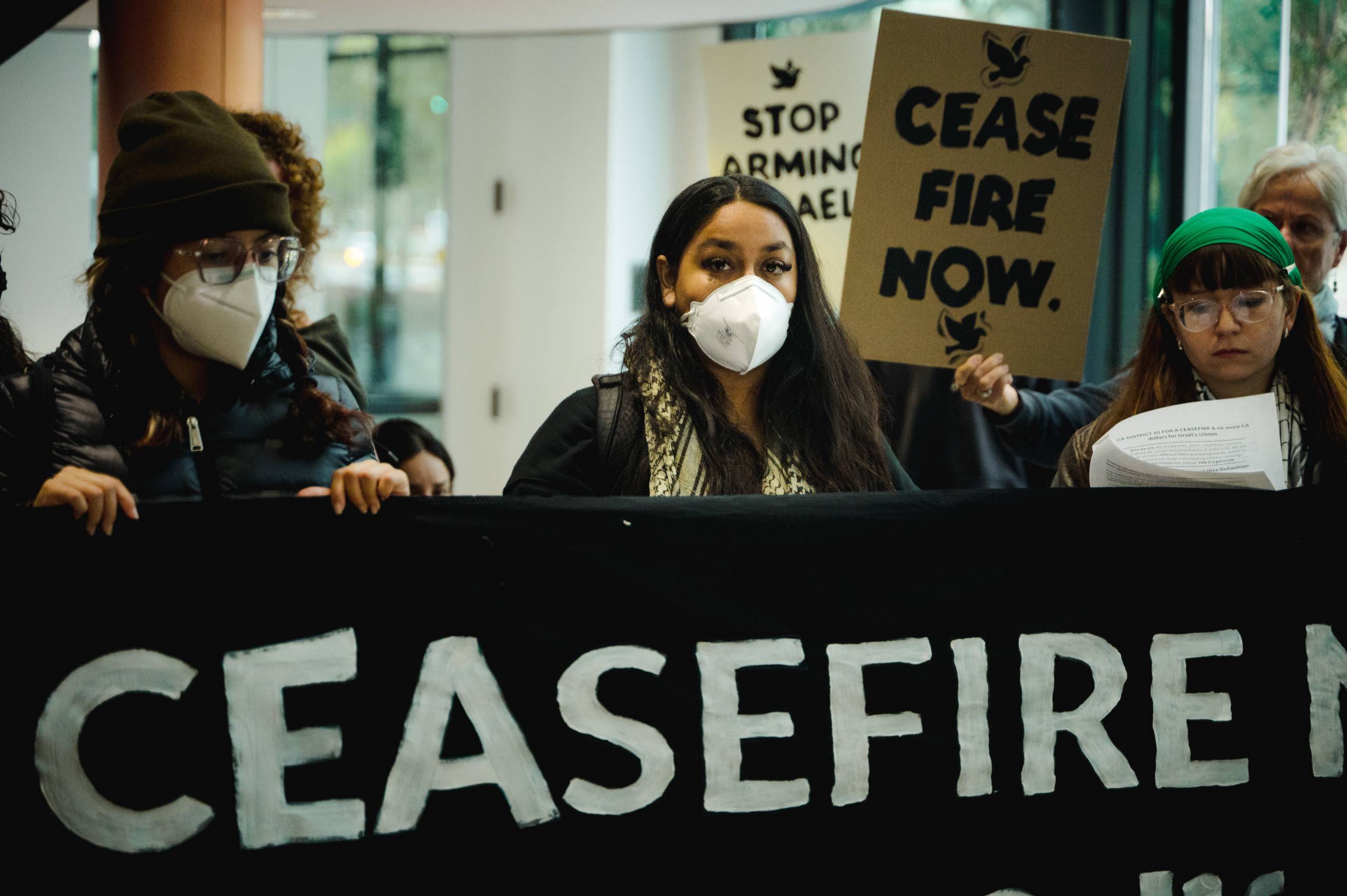 A woman with a white mask on stands in front of a black banner and infront of a sign that says cease fire now. 