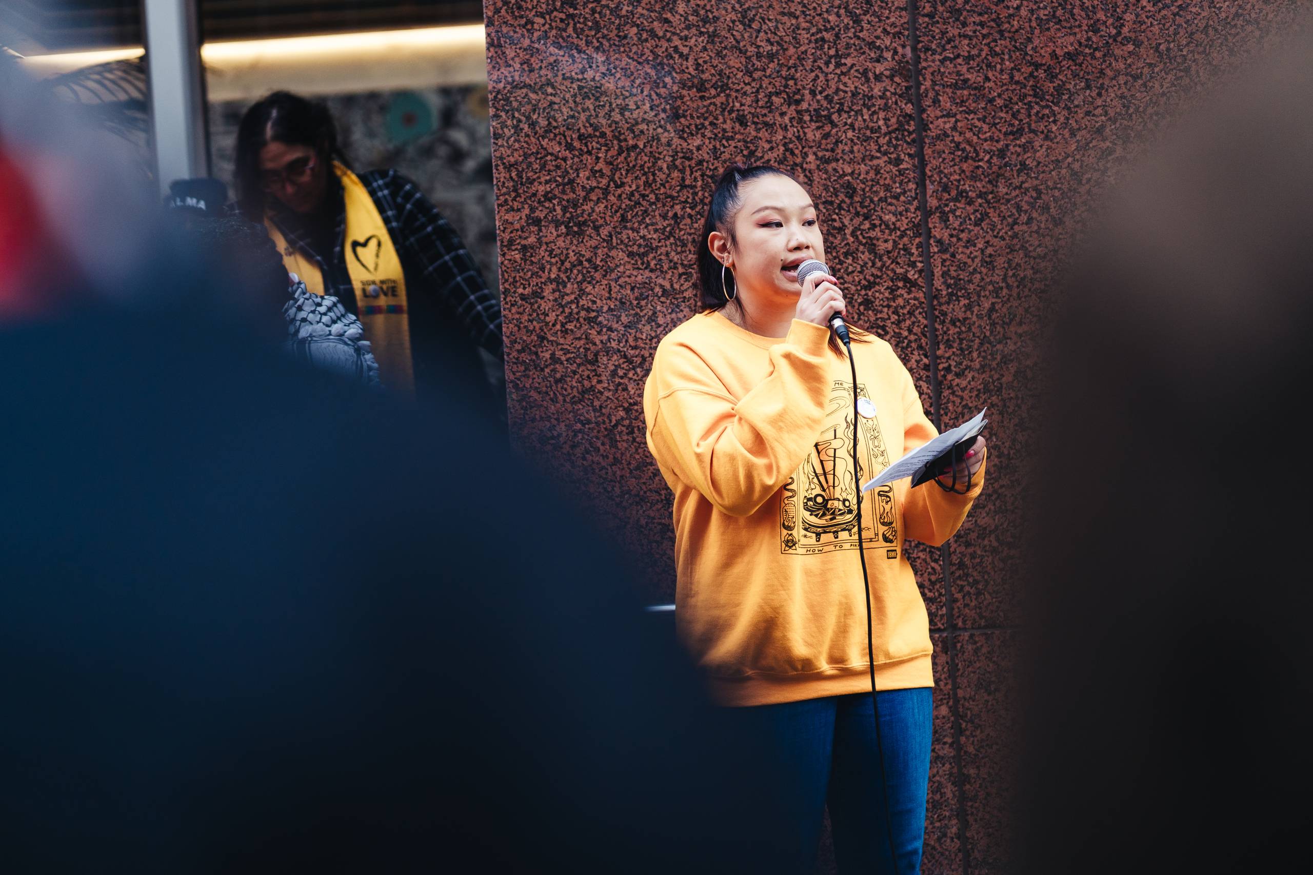 A woman holds a microphone and wears a yellow sweatshirt. 