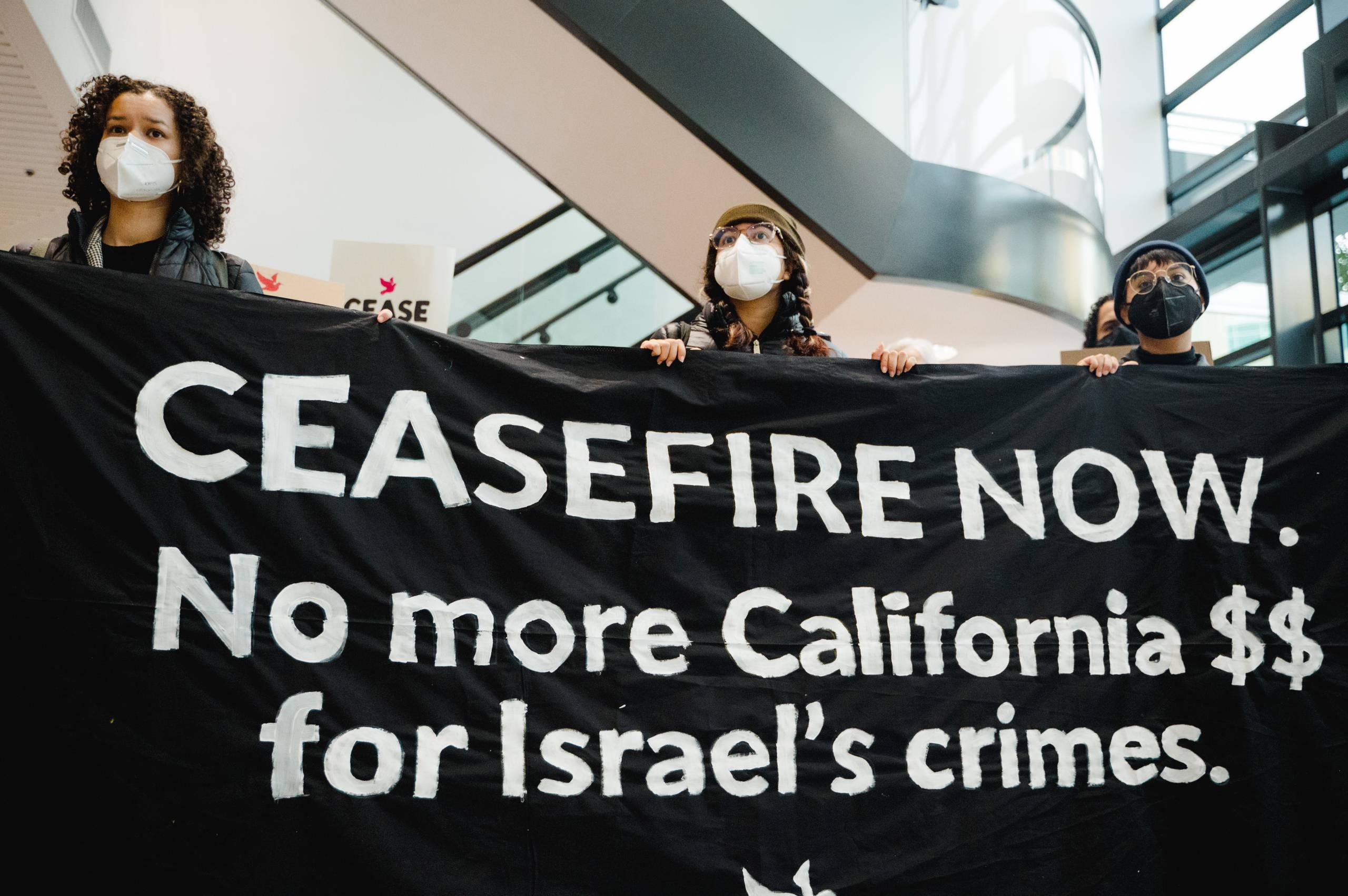 Three activists wearing white masks hold up a black banner that reads "No more California money for Israel's crimes." 