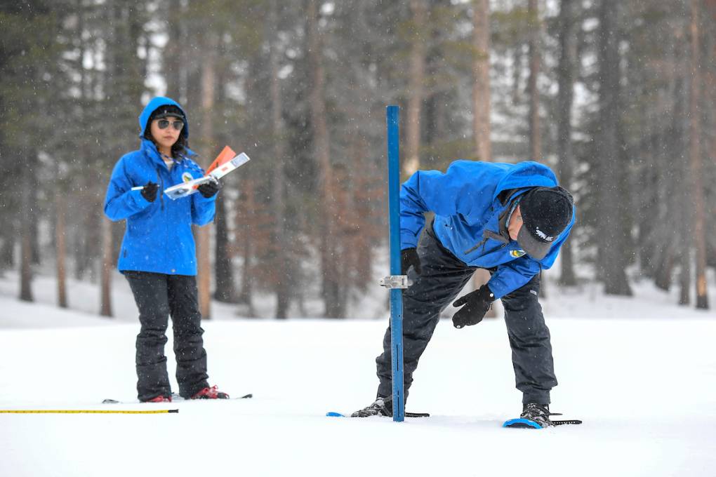 A man and woman dressed in blue snow jackets and black snow pants hold instruments. The man is bent over, measuring the snow.