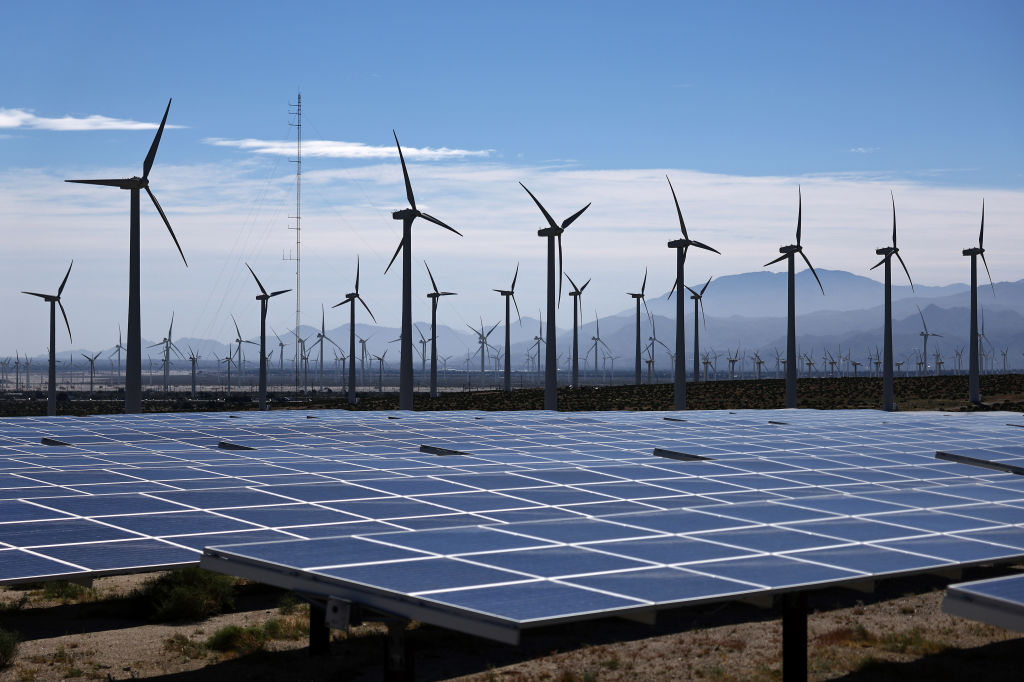 A wind farm full of wind turbines sits near solar panels beneath a somewhat cloudy blue sky.