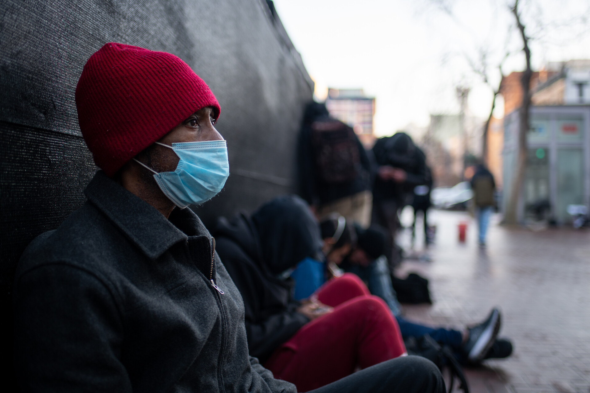 A close-up view of a man wearing a red beanie hat and a face mask with a dark jacket who is sitting next to other people on the sidewalk.