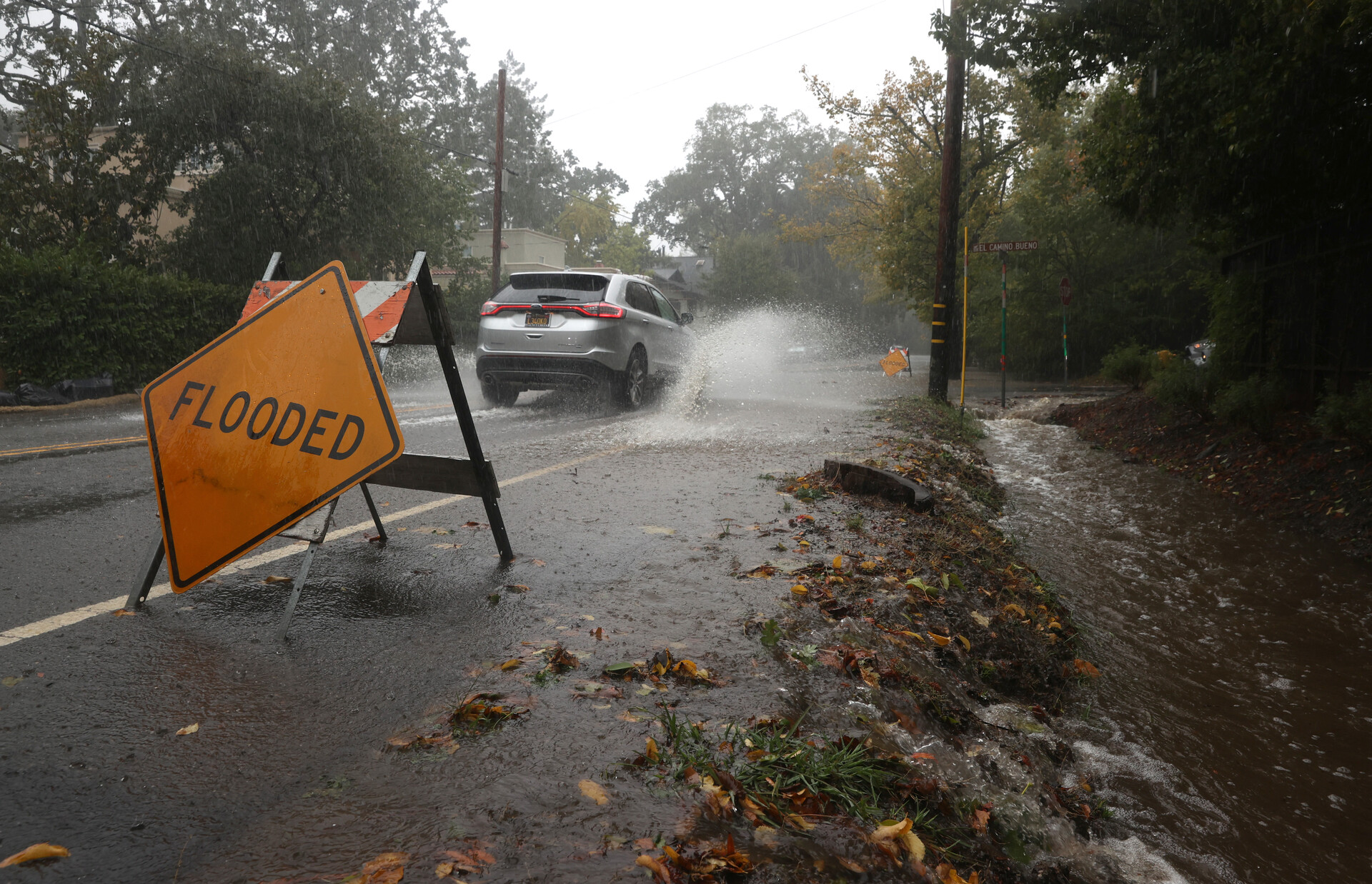 A car drives through water on a road, causing a splash with a sign that says 'Flooded' to the right.