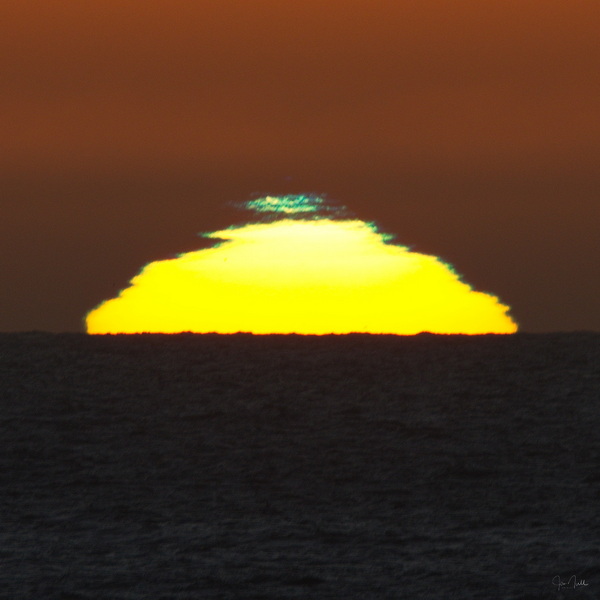 The yellow sun is seen setting into the horizon. A background of dark orange surrounds the sun. On the upper most part of the sun, you can see a faint green color.