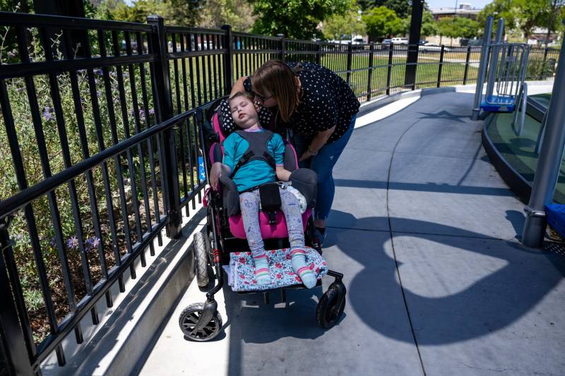 A woman leans over a young girl in a wheelchair on a sloping, curved outdoor concrete wheelchair ramp