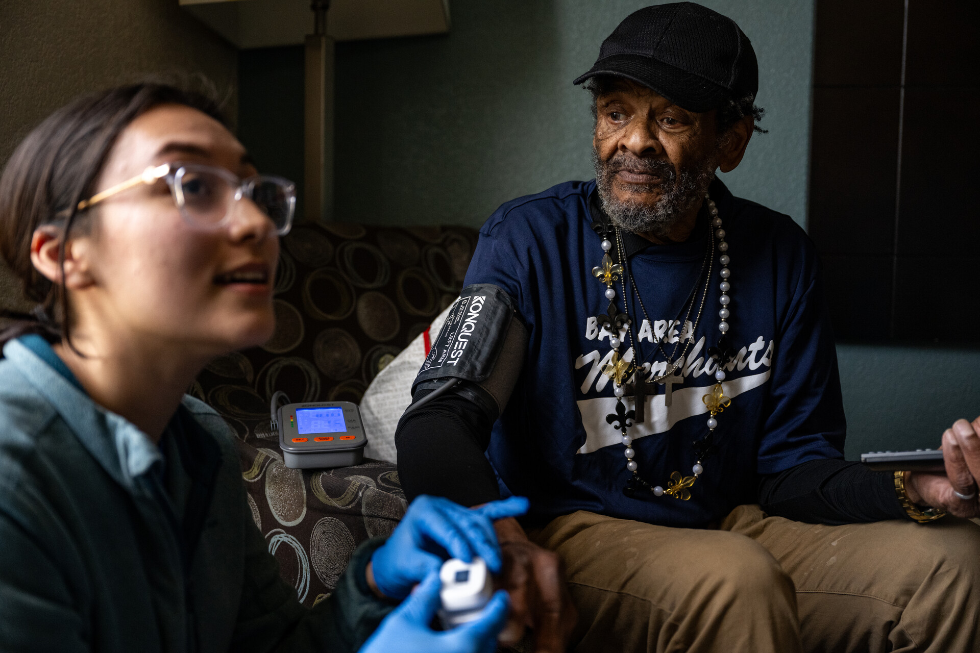 A woman smiling wearing blue protective nurse gloves sits in the foreground while an older man sits smiling at her while wearing a blood pressure monitor in the background