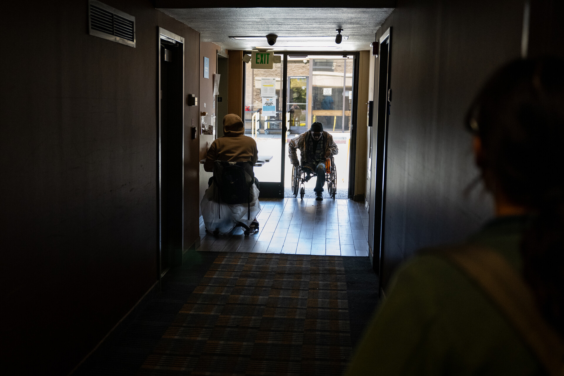 A man in a wheelchair enters a building from outside, seen in the distance backlit by daylight down a long, darkened hallway